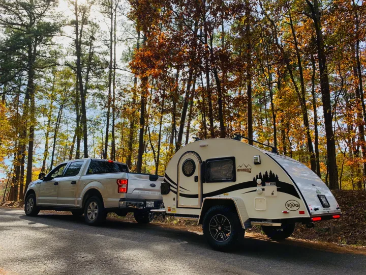 RV rental: A pickup truck tows a white teardrop camper on a sunny autumn road through a vibrant forest, ideal for camping adventures.. nuCamp T@G 2021