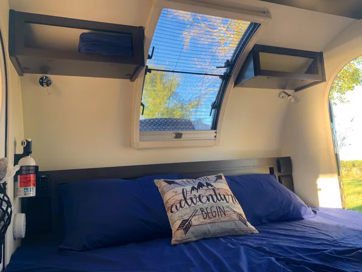 Cozy RV rental interior shows a blue bed, adventure pillow, overhead storage, & a bright skylight with a screen & outdoor view.. nuCamp T@G 2021