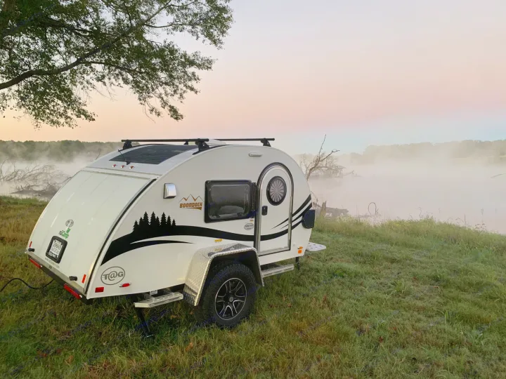 Misty sunrise scene: a white teardrop RV rental parked by foggy water, highlighting serene camping.. nuCamp T@G 2021