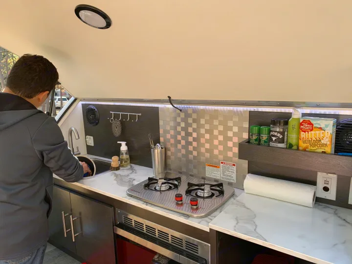 Man washes dishes in a compact RV rental kitchen with a stove and marble-look counters. Ideal for mobile living.. nuCamp T@G 2021