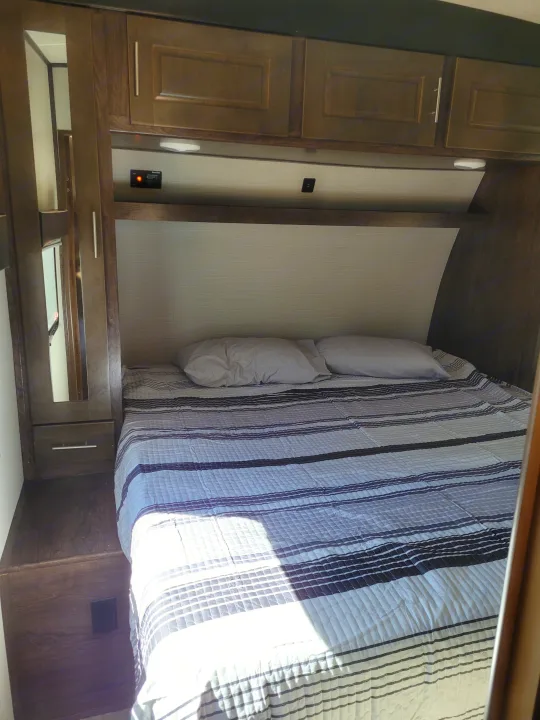 Inside an RV rental, a compact bedroom with a striped bed, pillows, overhead cabinets, and built-in storage.. Heartland Fuel 305 2018
