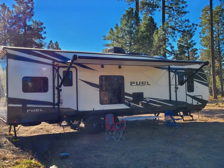 This Fuel travel trailer RV rental, parked at a sunny campsite under blue skies among tall pines, offers ultimate outdoor comfort.. Heartland Fuel 305 2018