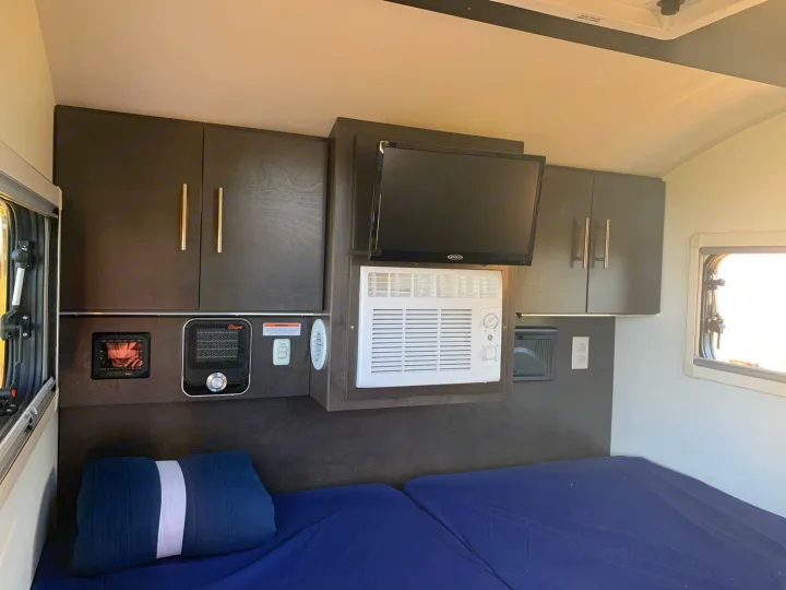 Inside an RV rental bedroom: dual beds, dark wood cabinets, a TV, window AC unit, and a heater for comfort.. nuCamp T@G 2021