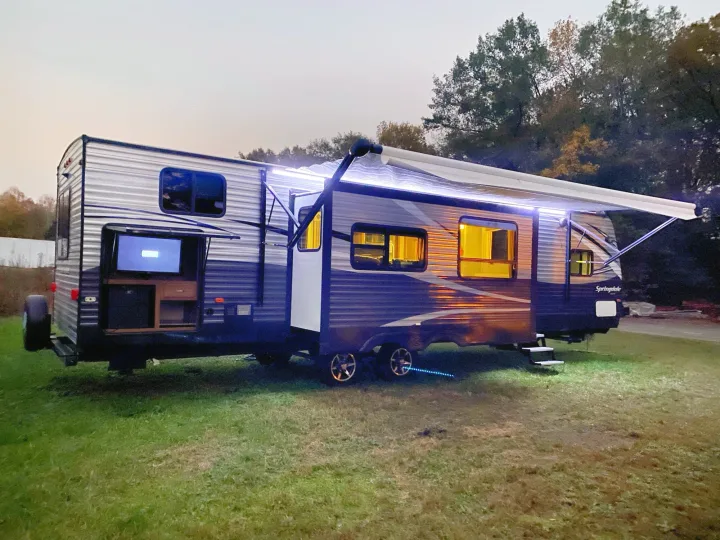 Evening glow of an RV rental: a modern travel trailer parked on grass, illuminated by interior, awning, and ground lights, with an outdoor TV.. keystone Springdale 2017