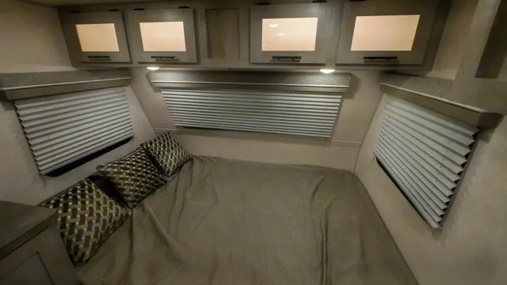 RV rental bedroom. Features a comfy bed with patterned pillows, pleated window blinds, and overhead cabinets for storage.. Forest River Ibex 2021