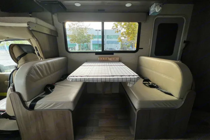 RV rental interior: A dinette with a checkered table, two upholstered benches (with seatbelts), and an overhead bunk.. Winnebago Spirit 2020