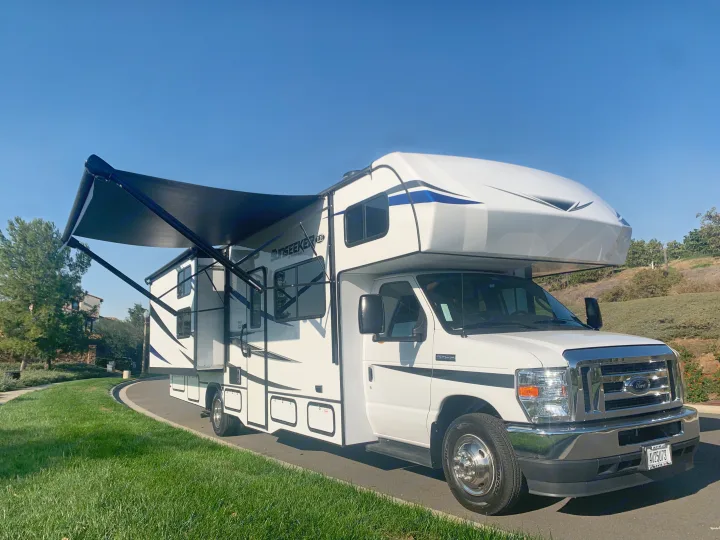 A white **rv rental** motorhome with its black awning extended, parked on an asphalt road beside lush green grass.. Forest River Sunseeker 2022