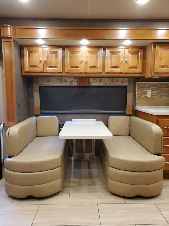 This RV rental interior features a cozy dining booth with beige seats, a white table, and light wooden cabinets.. Tiffin Motorhomes Allegro Open Road 2016