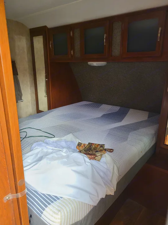 RV rental bedroom with a bed featuring a modern striped mattress, dark wood overhead cabinets, and a tall mirror.. Keystone Passport Ultra-lite Grand Touring 2016