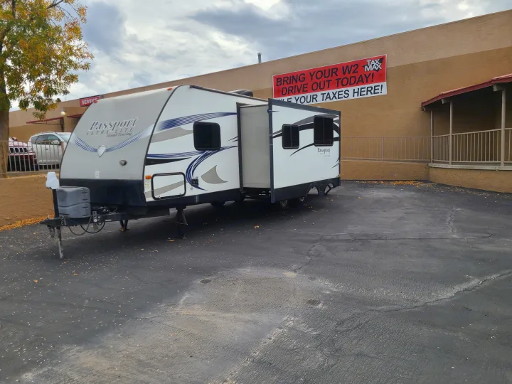 An extended Passport RV rental sits in a parking lot, with a "Tax Max" banner on the building behind it.. Keystone Passport Ultra-lite Grand Touring 2016