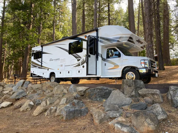 White Jayco Greyhawk RV rental parked on a leveled campsite, surrounded by tall trees and natural rocks.. Jayco Greyhawk 2021