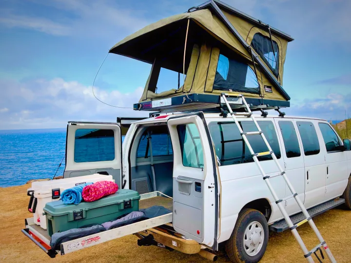 Adventure awaits with this RV rental! A white van with a deployed rooftop tent, ready for seaside camping & travel. Gear in back.. Ford Econoline 2009