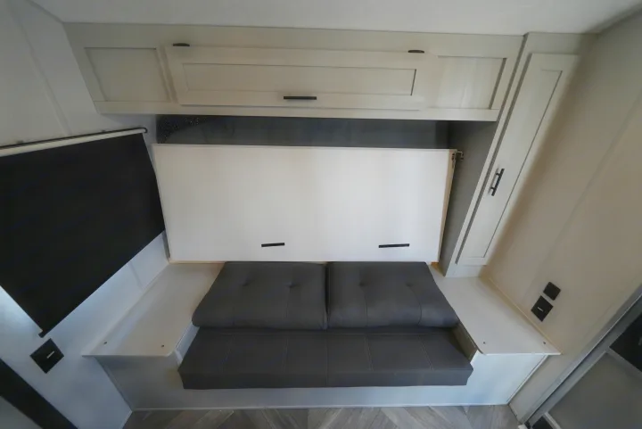 RV rental interior featuring a clever murphy bed setup with a gray sofa and overhead cabinets, maximizing space.. Forest River Wildwood FSX 2022