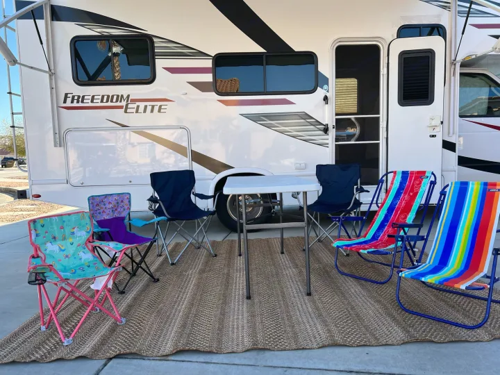 A white RV rental features an inviting outdoor setup: a woven rug, white table, and colorful chairs ready for relaxation.. Thor Freedom Elite 2020