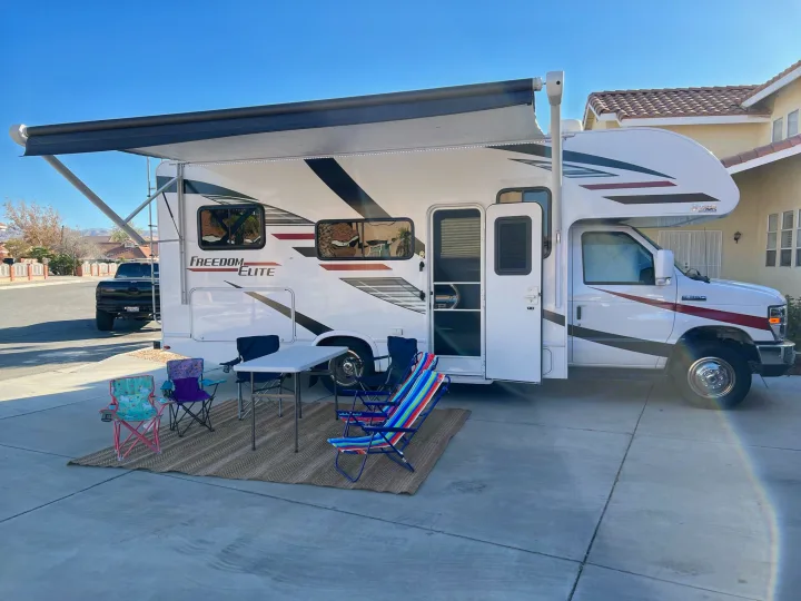 This white Freedom Elite rv rental is ready for outdoor relaxation, featuring an extended awning, mat, table, and colorful chairs.. Thor Freedom Elite 2020