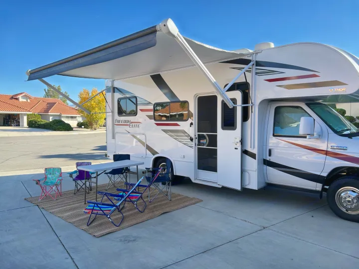 A white Thor Freedom Elite RV rental with an extended awning, outdoor table, & chairs on a mat, ready for camping fun.. Thor Freedom Elite 2020