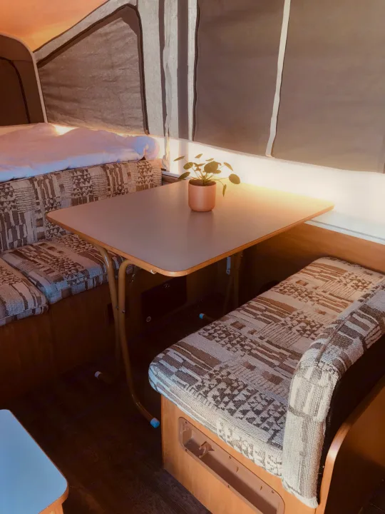 Cozy RV rental interior featuring a compact dinette, patterned benches, and a sleeping area with a white duvet.. Coleman Grand Tour Carmel 2003