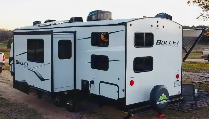 White Bullet Ultra Lite RV rental at a campsite with its slide-out extended, revealing windows and a power hookup.. Keystone Bullet 250BHS 2022
