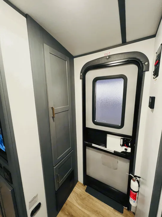 RV rental interior: Main entrance door with 'EXIT' sign, dark gray cabinets on left, fire extinguisher on right. Light wood floor.. Keystone Bullet 250BHS 2022
