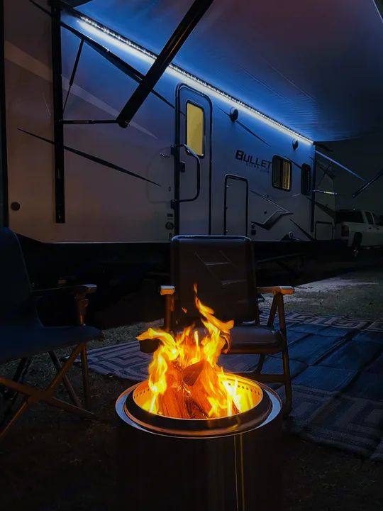 An inviting RV rental setup at night: glowing fire pit, comfy chairs, and brightly lit camper awning.. Keystone Bullet 250BHS 2022