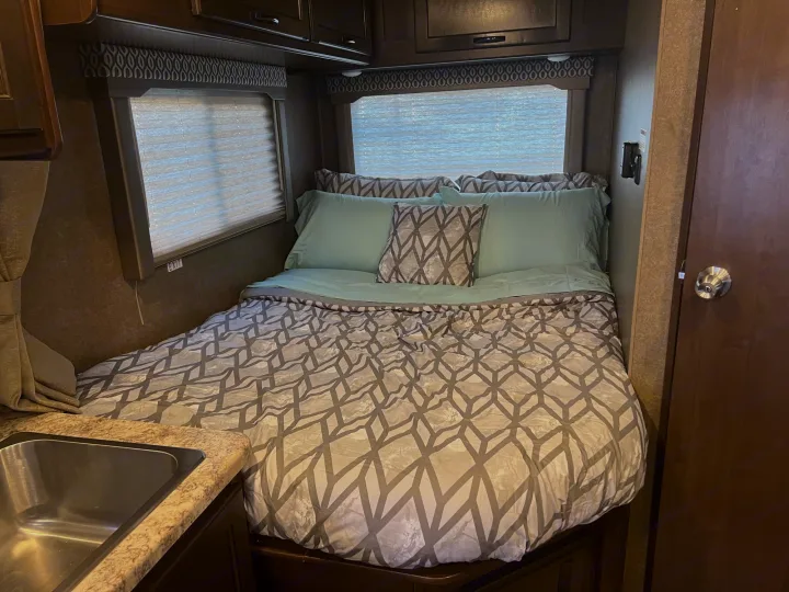Inside an RV rental: a comfortable bedroom with a geometric patterned comforter, blue sheets, and overhead storage.. Thor Freedom Elite 2020