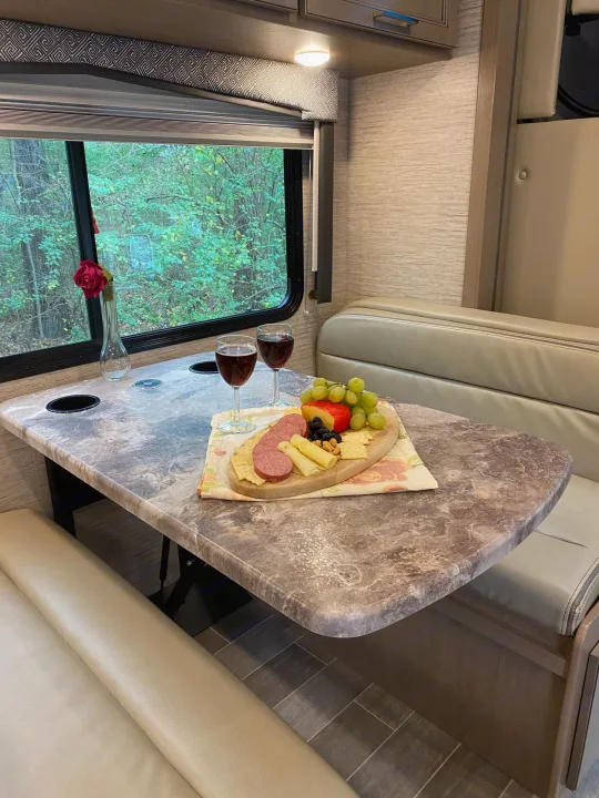 Cozy RV rental dinette with a charcuterie board, wine, and a red rose, offering a scenic view of lush trees outside.. Thor Magnitude GA 2022