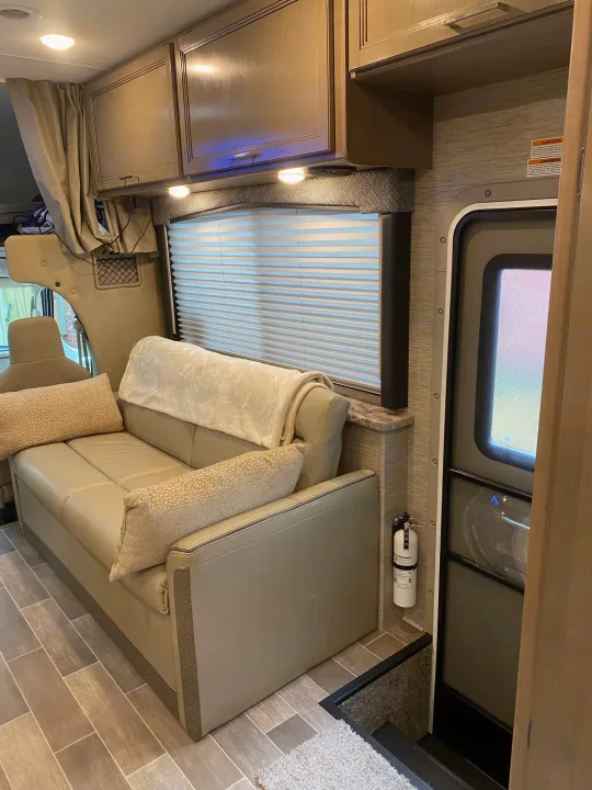Inside a modern **RV rental**: view of tan sofa, overhead storage, window blinds, and entry door with fire extinguisher.. Thor Magnitude GA 2022