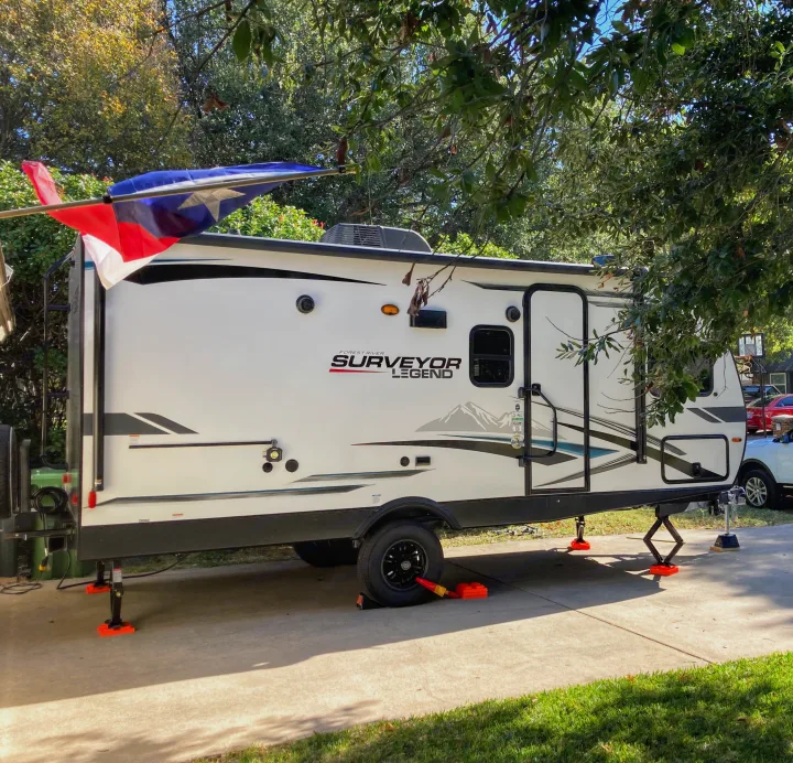 A white Surveyor Legend travel trailer, stabilized on concrete, parked under trees, proudly flying a Texas flag. Ideal for your RV rental getaway.. Forest River Surveyor Legend 2022