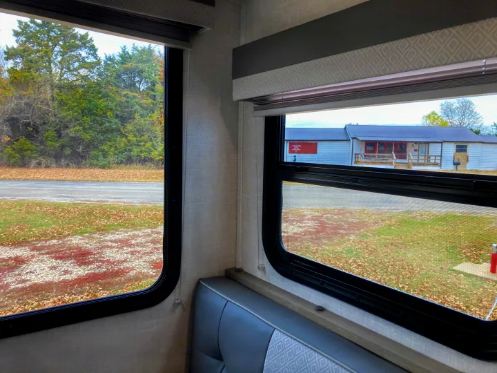 RV rental interior. A gray bench seat and windows frame an autumn outdoor view of trees, a road, and a distant house.. Forest River Surveyor Legend 2022