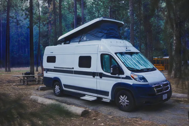 A white pop-top RV rental camper van parked on pavement in a dense forest campsite. A picnic table is nearby.. Hymer Hymer Class B 2019