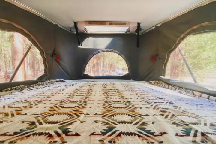 Inside an RV rental: a spacious pop-top bed with a Southwestern blanket and mesh windows overlooking a forest campsite.. Hymer Hymer Class B 2019