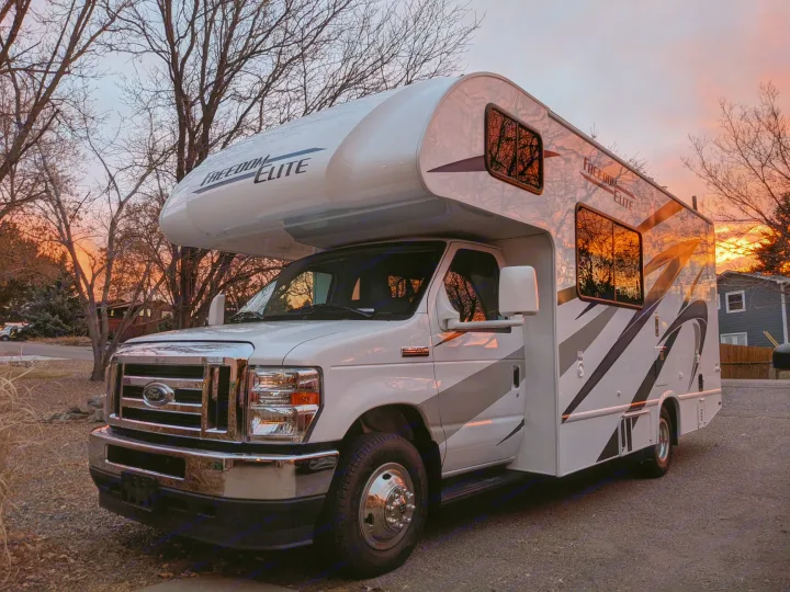 White Ford Freedom Elite Class C RV rental reflects vibrant sunset colors and bare trees in a driveway.. Thor Freedom Elite 2021