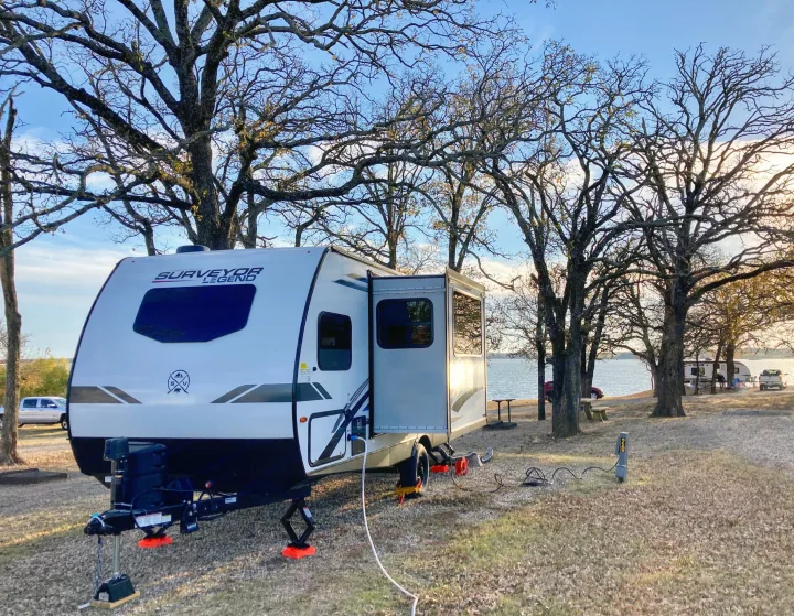 Lakeside rv rental: A white travel trailer is set up with power and slide-out extended, under bare trees by a lake.. Forest River Surveyor Legend 2022