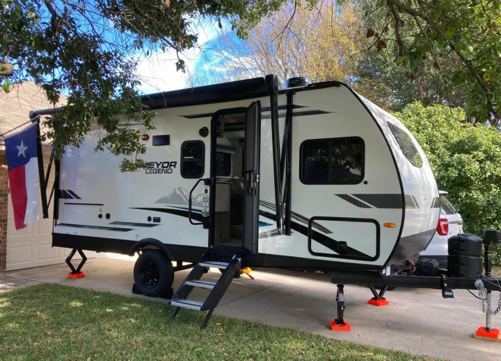 A white Surveyor Legend RV rental is parked on a driveway, with a Texas flag hanging from its side, under trees.. Forest River Surveyor Legend 2022