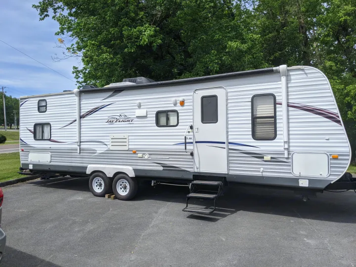 This white and gray Jay Flight travel trailer, an excellent RV rental option, is parked outdoors with green trees in the background.. Jayco Jay Flight 2012