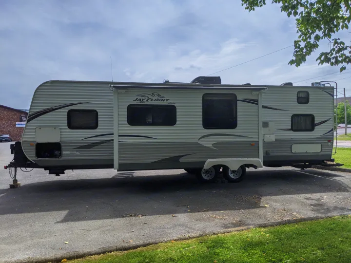 A white and grey Jayco Jay Flight travel trailer, an RV rental, parked on asphalt under a partly cloudy sky. Awning extended.. Jayco Jay Flight 2012