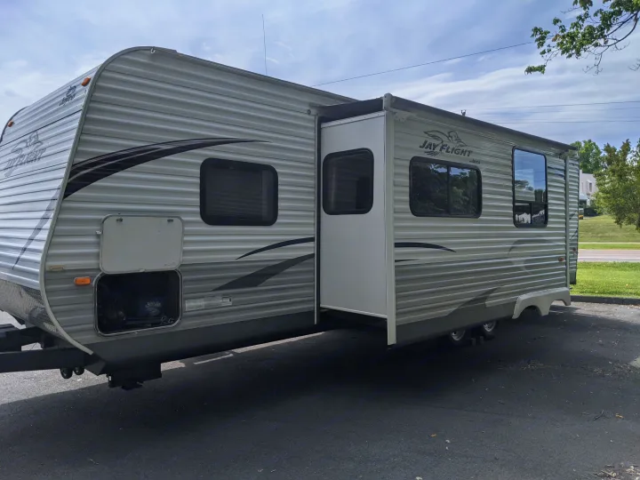 A grey Jay Flight travel trailer with an extended slide-out, available for RV rental, parked on asphalt under a cloudy sky.. Jayco Jay Flight 2012
