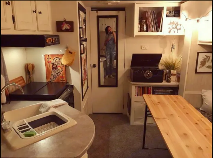 View inside an RV rental: kitchen sink, stove, record player, and a wooden table beside a bed. Person reflected in mirror.. Heartland North Trail 2009