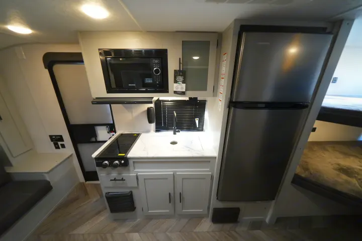 Interior of a modern RV rental, featuring a compact kitchen with microwave, sink, stove, and fridge, alongside a dinette and bed.. Wildwood FSX 178BHsk 2022