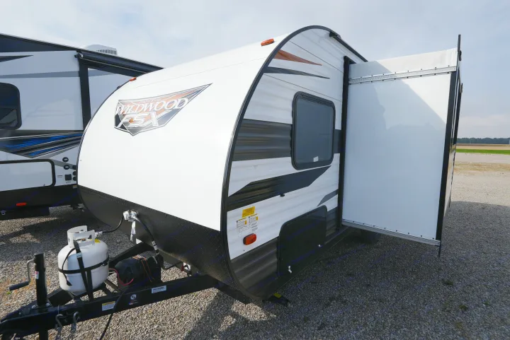 A white Wildwood FSX travel trailer with black and brown accents, showing its extended side, on a gravel lot. Ideal for RV rental.. Wildwood FSX 178BHSK 2022