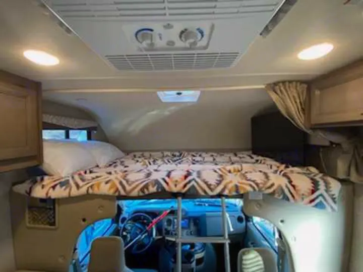 Comfortable cab-over bunk in an RV rental, featuring a bed, overhead AC, and view into the driver's cockpit.. Thor Freedom Elite 2021