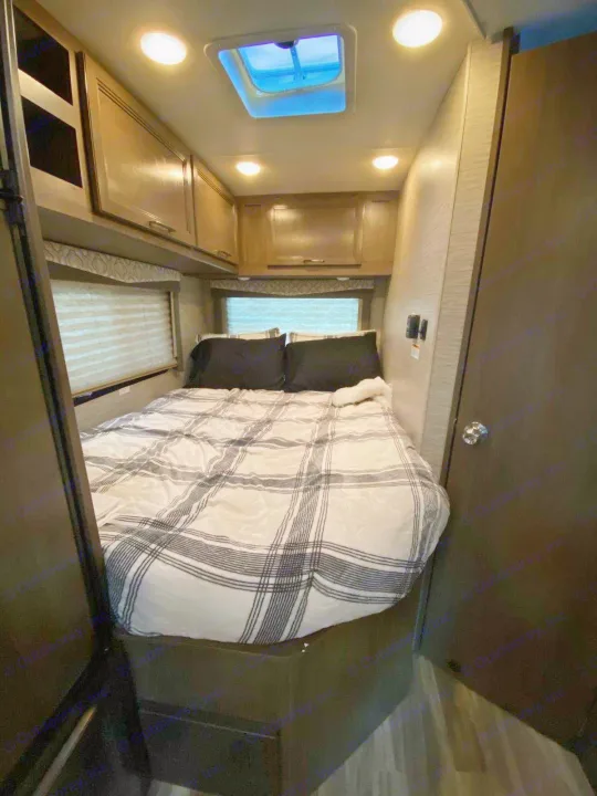 A cozy RV rental bedroom features a plaid bed, overhead wood-tone cabinets, and bright ceiling lights with a skylight.. Thor Freedom Elite 2021