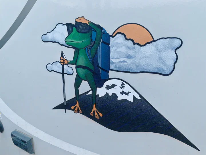 A playful decal of a frog hiker on an RV rental, sporting sunglasses and a backpack on a snowy mountain peak.. Forest River R-Pod 2020