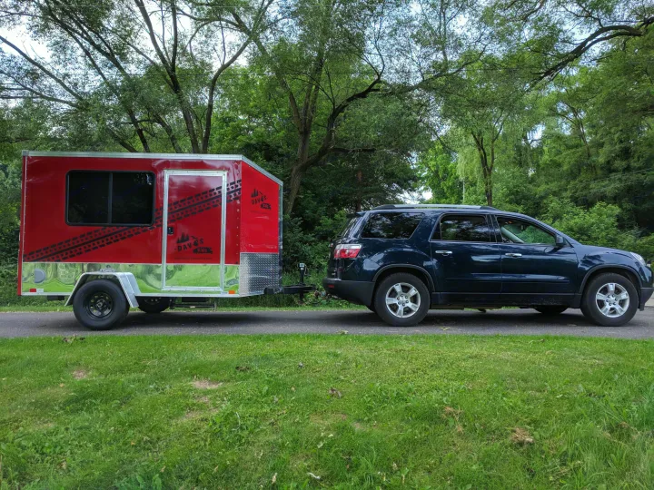A dark SUV tows a red and silver Dave's RV rental trailer on an asphalt path amidst green trees.. United XLMTV-610 2018