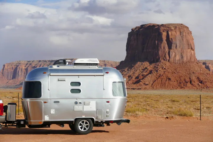 Silver Airstream travel trailer (RV rental) parked on red dirt, framed by towering red rock mesas under cloudy skies.. Airstream Caravel 2020