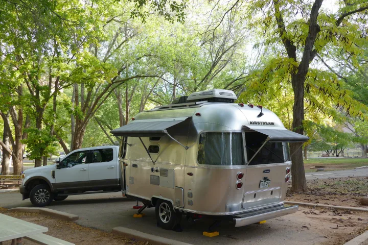 An Airstream RV rental, with awnings extended, is hitched to a white truck at a scenic, tree-filled campsite.. Airstream Caravel 2020