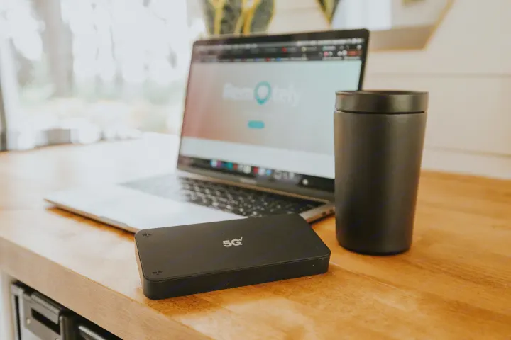 A modern remote workspace on a wooden table, featuring a laptop, 5G hotspot, and travel mug, ideal for RV rental journeys.. Ram Promaster 2500 2021