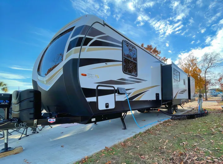 A white and black RV rental travel trailer is parked at a sunny campsite, connected to utilities.. Keystone RV Outback 2021