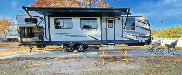 This large white travel trailer RV rental is parked at a fall campsite with an outdoor kitchen, picnic table, and chairs.. Keystone RV Outback 2021
