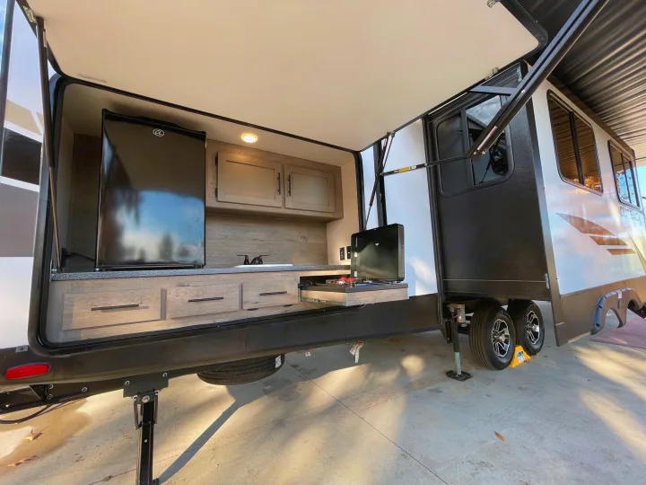 RV rental's outdoor kitchen with fridge, sink, pull-out stove, and cabinets. Includes leveling jacks and wheels for stability.. Keystone RV Outback 2021